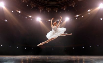 A ballet dancer in white tutu leaping under lights on stage. Adobe